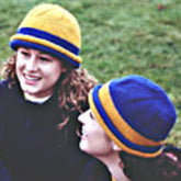 Two people are outdoors, both wearing Bryson Distributing, Inc.'s School Colors Hat made of beginner-friendly yarn. They are smiling and appear to be enjoying a lively conversation. One School Colors Hat features a yellow top with blue bands, while the other has a blue top with yellow bands. A grassy background is visible.
