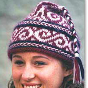 A person is smiling and wearing a beautiful Swirls Hat from Bryson Distributing, Inc., showcasing a red, white, and purple pattern. The knitted Swirls Hat features intricate designs and has a braided tassel hanging from the top. The background is blurred, emphasizing the person and their hat.