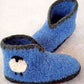 The Crocheted Boot Slipper Pattern by Bryson Distributing, Inc. features a pair of blue, fuzzy baby booties adorned with dark grey trim around the tops and bottoms. Each bootie is charmingly decorated with a small, white sheep design on the side, combining slipper-like comfort with adorable charm.