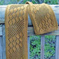 The Streaming Leaves Scarf by Bryson Distributing, Inc. drapes elegantly over a wooden rail. Crafted in a knitted golden brown, it showcases intricate diamond-shaped openwork patterns reminiscent of leaf lace interspersed with solid knitted sections. Greenery is partially visible through the wooden rail in the background, adding to its serene charm. This project is perfect for an adventurous beginner.