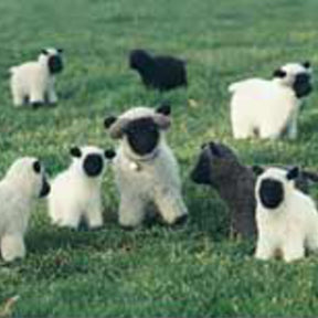 Bryson Distributing, Inc.'s "A Felt Flock (sheep)" features plush toy lambs, some white and some black, scattered across a lush green lawn. The toys have black faces and hooves, with one prominent gray lamb in the center. Featuring felted knitting and natural colors, the scene mimics a flock of lambs grazing in the field.