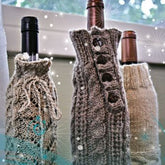 Three Wine Bottle Cozies from Bryson Distributing, Inc. are wrapped in cozy, stylish knitted covers, each uniquely designed with different patterns and buttons. They stand on a window sill with a blurred background featuring a green outdoor scenery and delicate light effects resembling sparkles, making for an elegant wine gift wrap.