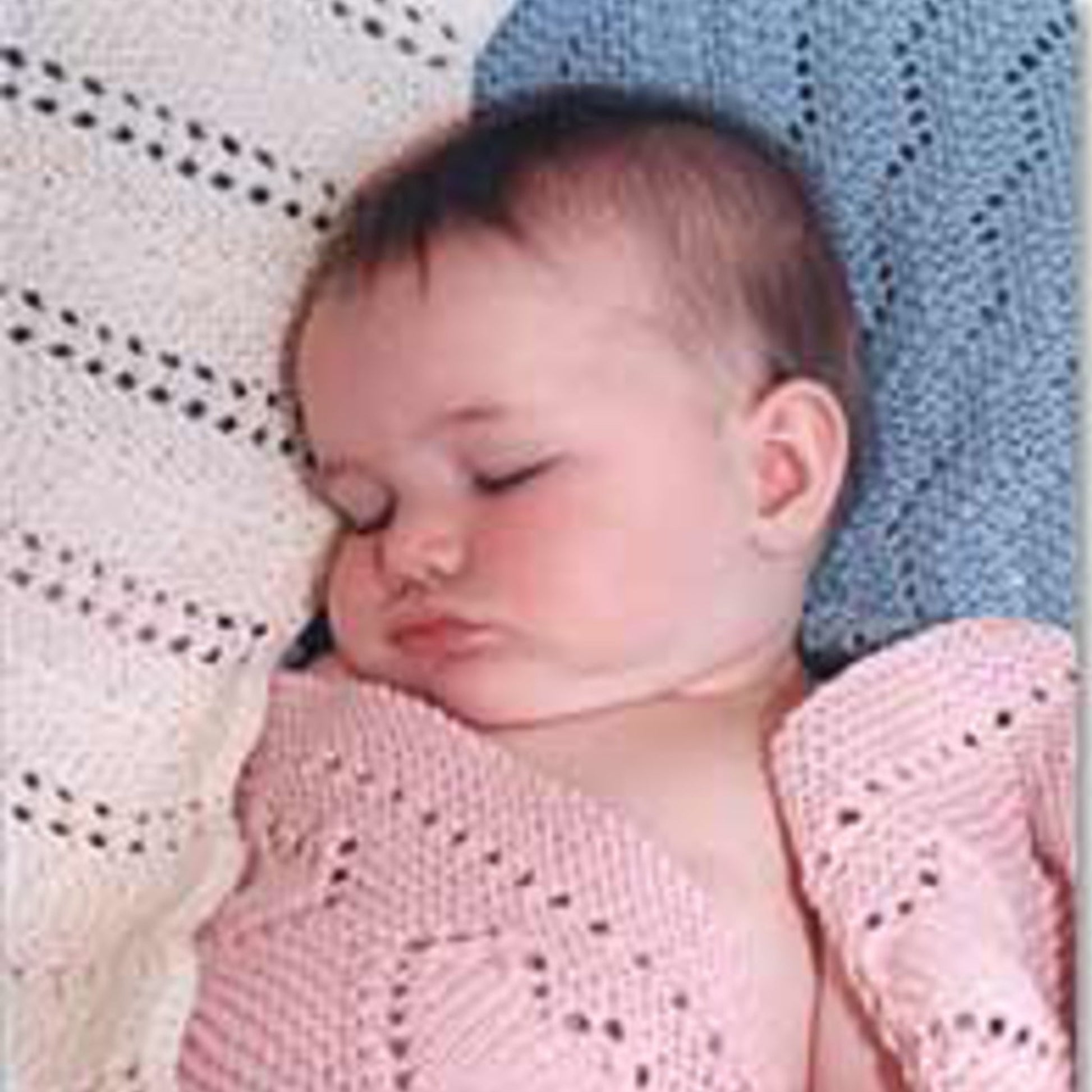 A baby peacefully sleeps on a blanket from the Easy Knit Baby Blankets Collection 2 by Bryson Distributing, Inc. The baby is cozily wrapped in a pink blanket and rests on a blue and white background adorned with beautiful knitting patterns. With closed eyes and slightly puckered lips, the baby's serene and calm expression shines during nap time.