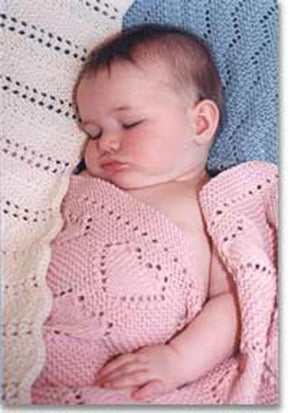 A baby peacefully sleeping, wrapped in a pink crocheted blanket with heart patterns from the Easy Knit Baby Blankets Collection 2 by Bryson Distributing, Inc. The baby lies on a soft, textured blanket that is white and blue, crafted from worsted weight yarn. The baby's eyes are closed, and their cheeks are slightly rosy.