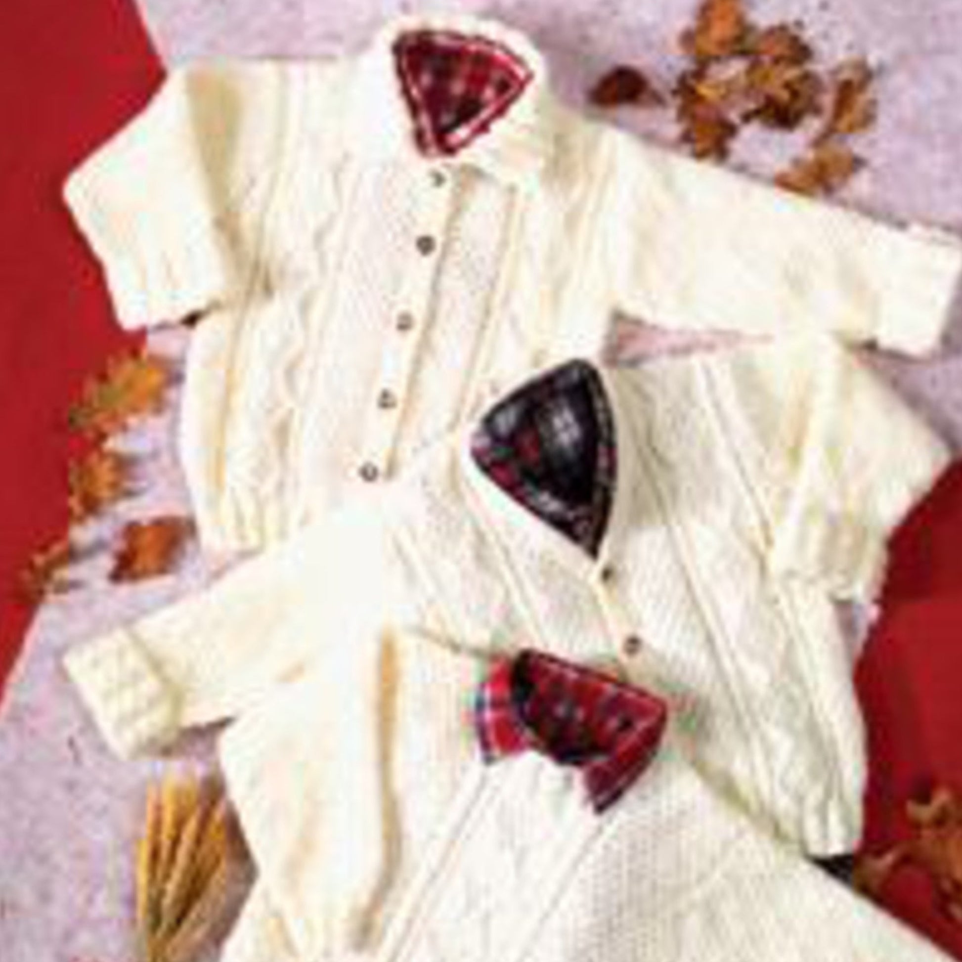 Two Encore Children's Sweater & Jacket pieces from Plymouth Yarn Co, featuring cream-colored cabled designs with button-down fronts and plaid linings, are displayed on a red and white surface. Autumn leaves and wheat stalks scattered around the garments add a seasonal touch, making them perfect for kids during the fall.