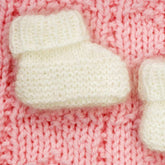 A pair of white Angora Baby Booties from Plymouth Yarn Co, perfect for preemie to newborn sizes, sit on a knitted pink background. The intricate stitches of both the booties and the background are clearly visible, highlighting their soft and cozy texture.