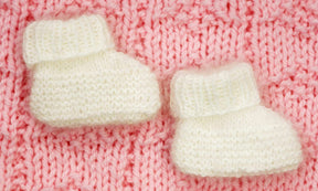 Two small Angora Baby Booties by Plymouth Yarn Co are placed side by side on a textured, pink knitted fabric. The cozy design features folded cuffs at the top, perfect for preemie to newborn sizes.