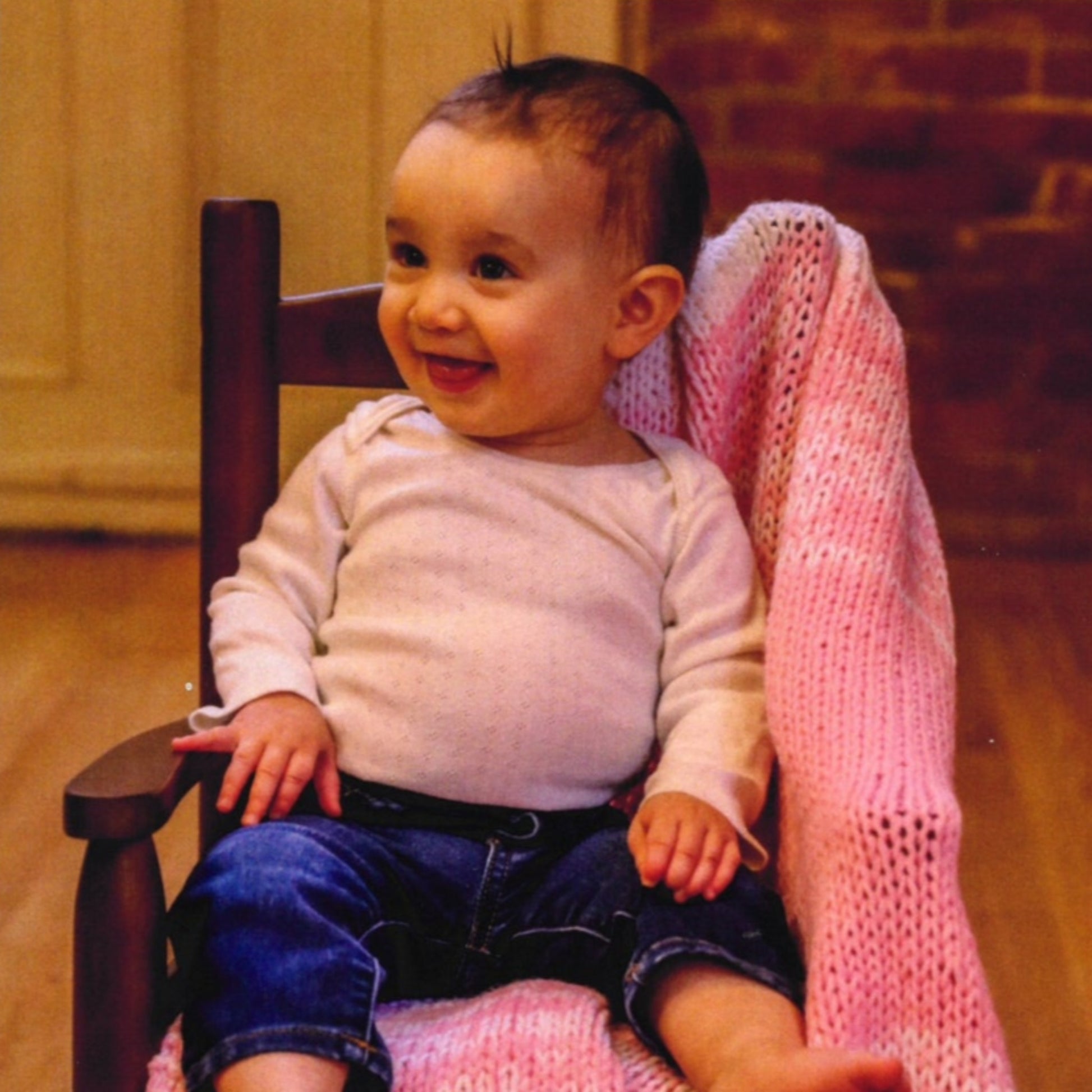 A baby with short dark hair sits on "The Encore 8-Hour Baby Blanket Revisited" by Plymouth Yarn Co, handwoven from soft yarn, draped over a wooden chair. They are wearing a white long-sleeve top and blue jeans, smiling and looking to the side. The background features a brick wall and wooden floor.