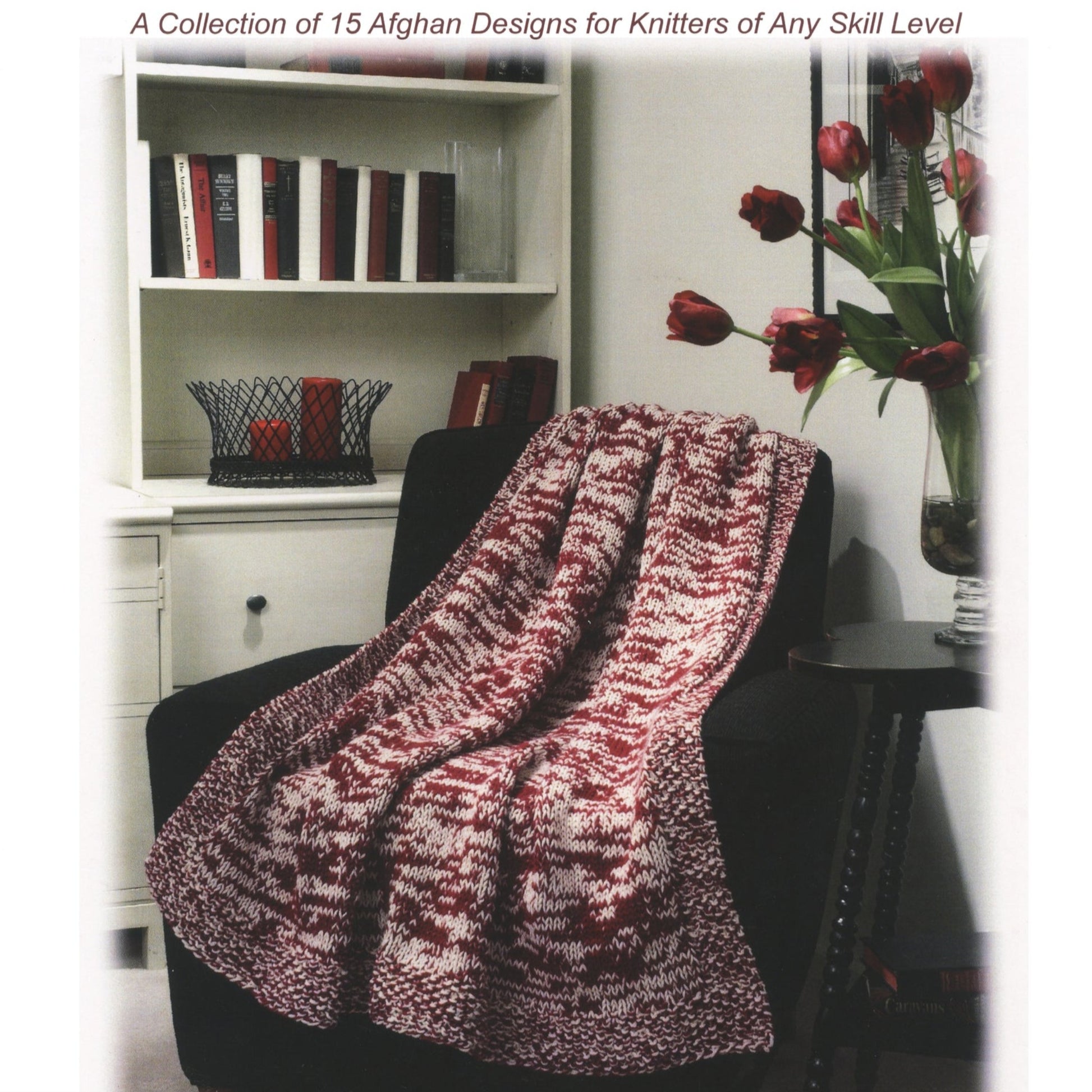 A cozy living room corner featuring a black armchair draped with a Done By Monday Afghan by Plymouth Yarn Co, showcasing a beautiful red and white pattern. A white side table with a flower vase sits beside the chair. In the background, a bookshelf holds books and a decorative basket, offering perfect inspiration for knitters' afghan designs.