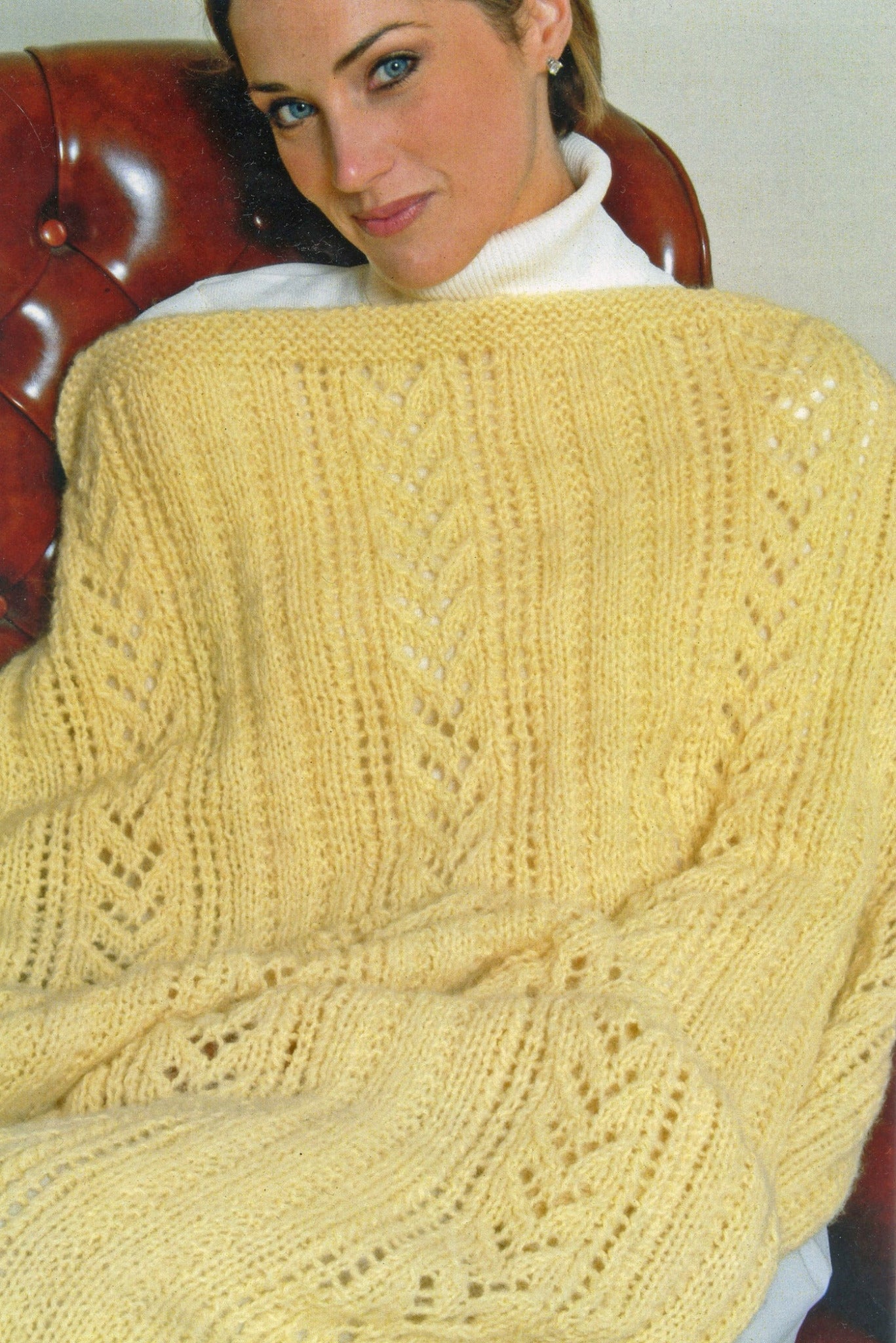 A person is seated on a red leather chair, wearing a white turtleneck and wrapped in the Plymouth Yarn Co's Lace & Rib Afghan, a handmade yellow knitted afghan featuring intricate patterns. They are smiling softly and looking at the camera. The cozy, textured blanket made from bulky yarn is the focus of the image.