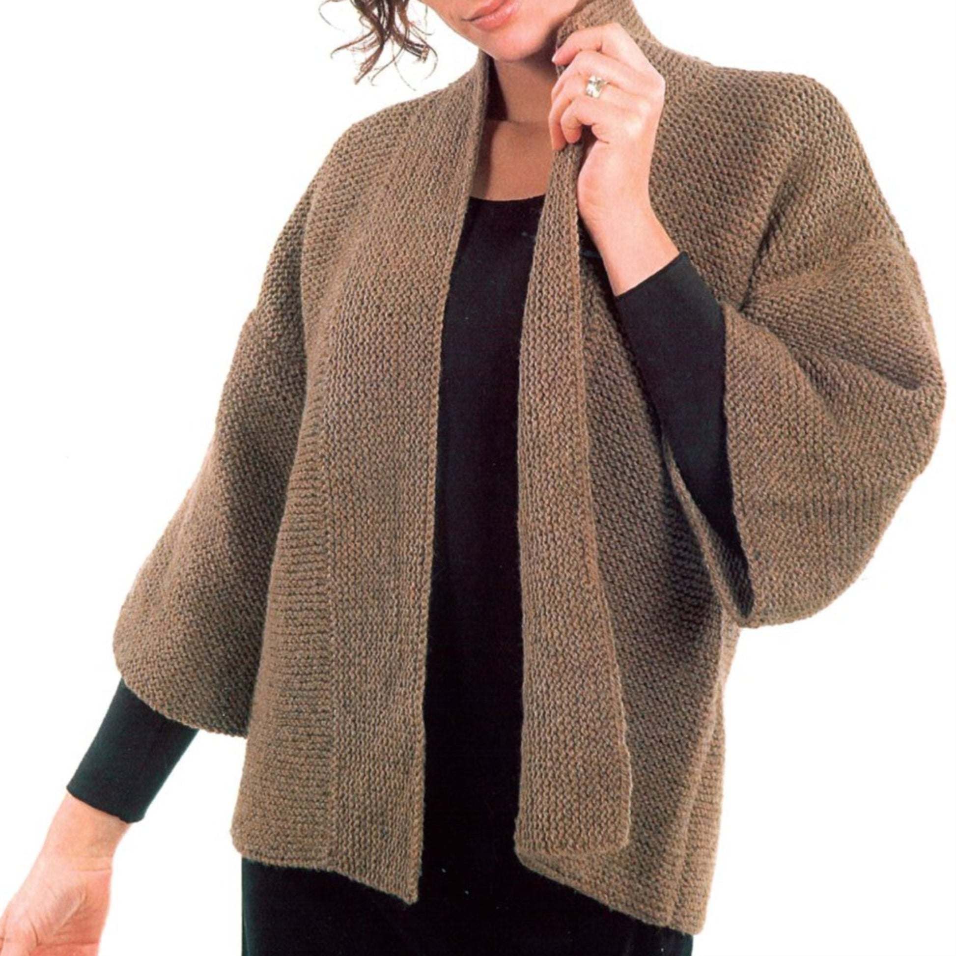 A person is wearing a loose-knit, brown cardigan with wide sleeves over a black shirt. The Basic Garter Stitch Kimono by Plymouth Yarn Co has a shawl collar and a textured pattern reminiscent of garter stitch kimono designs. The person is holding the collar with one hand, their face partially out of the frame.