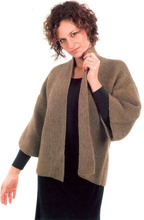 A person with curly hair is wearing the Basic Garter Stitch Kimono, a brown knitted cardigan from Plymouth Yarn Co, over a black long-sleeved outfit made from worsted weight yarn. The kimono has wide sleeves and an open front, and the individual has a slight smile while holding it near the neck with one hand.