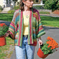 A woman carrying two pots of orange chrysanthemums walks along a paved path. She wears a Basic Garter Stitch Kimono by Plymouth Yarn Co over a yellow shirt and blue jeans. Green trees and a house are visible in the background.