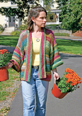 A woman carrying two pots of orange chrysanthemums walks along a paved path. She wears a Basic Garter Stitch Kimono by Plymouth Yarn Co over a yellow shirt and blue jeans. Green trees and a house are visible in the background.