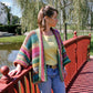 A woman stands on a red wooden bridge near a serene pond, looking to the side. She is adorned in Plymouth Yarn Co's Basic Garter Stitch Kimono, a colorful, striped knitted cardigan made from worsted weight yarn, layered over a yellow top and blue jeans. In the background, lush greenery and buildings evoke a tranquil park setting.