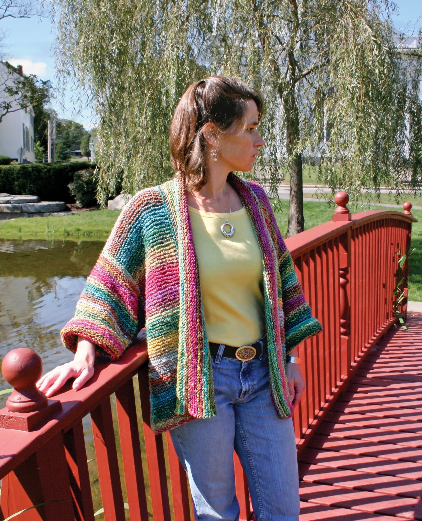 A woman stands on a red wooden bridge near a serene pond, looking to the side. She is adorned in Plymouth Yarn Co's Basic Garter Stitch Kimono, a colorful, striped knitted cardigan made from worsted weight yarn, layered over a yellow top and blue jeans. In the background, lush greenery and buildings evoke a tranquil park setting.