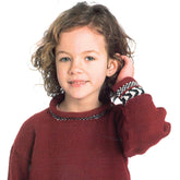 A young child with wavy, medium-length hair smiles slightly while touching their hair with one hand. They wear a red Plymouth Yarn Co Child's Fair Isle Pullover featuring black and white trim at the cuffs against a plain white background.