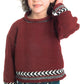 A young child with curly hair is wearing a Plymouth Yarn Co Child's Fair Isle Pullover, featuring intricate trims on the neck, sleeves, and hem. The cozy red pullover is paired with blue jeans. The child has a slight smile and one hand touching their head against a plain white background.