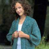 A young girl with curly hair stands outdoors, wearing a Plymouth Yarn Co Kid's Kimono in teal over a white top that mimics kimono styling. She gazes at the camera with a slight smile, holding the edges of the piece. The background is blurred greenery.