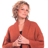 A woman with curly blonde hair is wearing a rust-colored Kimono Stockinette Stitch with Garter Border by Plymouth Yarn Co, layered over a black top. The ensemble looks comfortable to wear as she smiles slightly, holding her hands together in front of her. The background is white and uncluttered.