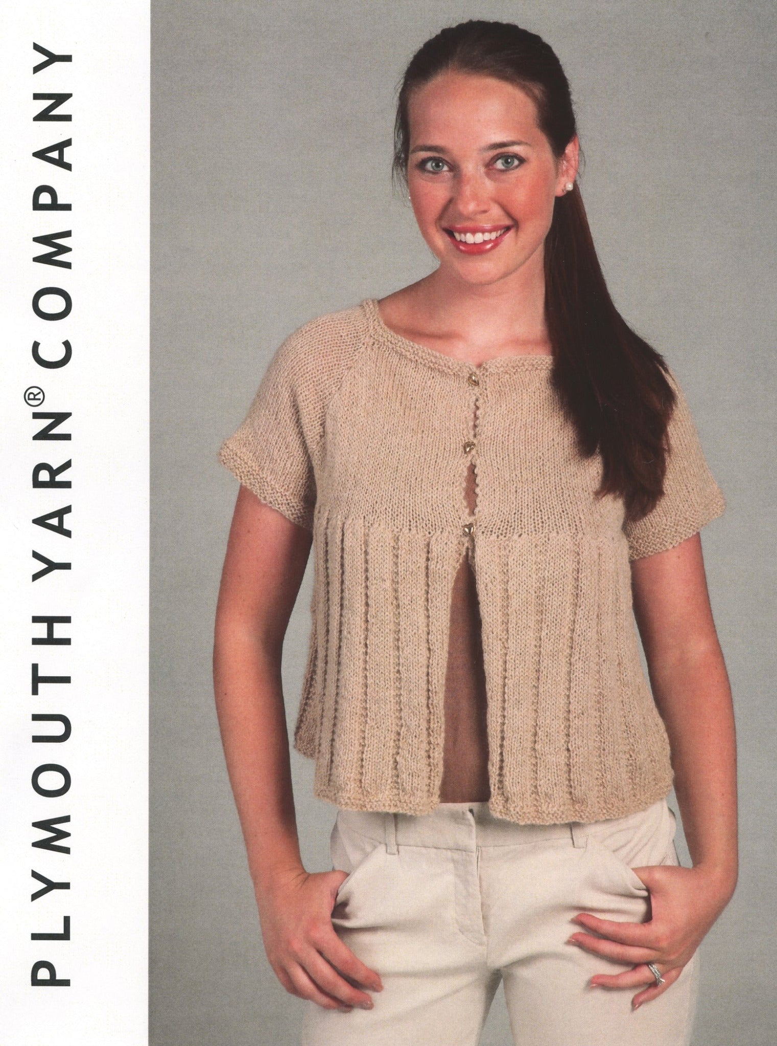 A woman is seen wearing a stylish Loose Knit Swing Cardigan in beige, hand-knit with Alpaca yarn, featuring short sleeves and pleated details. The cardigan is open at the front and fastened with a single button near the top. She pairs it elegantly with white pants. On the left, text reads "Plymouth Yarn Co." She stands against a neutral background.