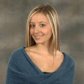 A young woman with straight, blonde hair and light skin smiles at the camera, wrapped in a soft blue Mobius Capelet by Plymouth Yarn Co, exposing her shoulders and upper chest. The background is a gray, mottled texture.