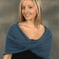 A young woman with long blonde hair stands in front of a neutral backdrop, smiling at the camera. She is wearing a soft, black top underneath the blue knitted Mobius Capelet from Plymouth Yarn Co, which drapes over her shoulders and crosses in the front—an easy-to-knit design available for download.