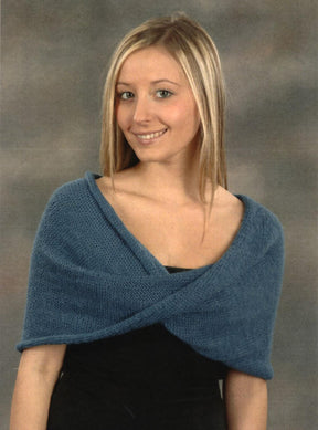 A young woman with long blonde hair stands in front of a neutral backdrop, smiling at the camera. She is wearing a soft, black top underneath the blue knitted Mobius Capelet from Plymouth Yarn Co, which drapes over her shoulders and crosses in the front—an easy-to-knit design available for download.