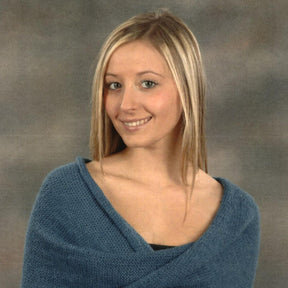 A young woman with long blonde hair is smiling at the camera. She is wearing a soft blue shawl from Plymouth Yarn Co, wrapped around her shoulders against a gray, textured background. The shawl, named "Mobius Capelet," adds an elegant touch to her outfit.