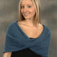 A young woman with long, straight blonde hair is smiling at the camera. She is wearing a dark top with an elegant Mobius Capelet in soft blue, draped over her shoulders from Plymouth Yarn Co. The background features a neutral, grayish-brown gradient.