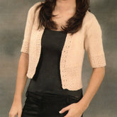 A woman wearing a black tank top and a stylish Wrap Cardigan by Plymouth Yarn Co, designed in light beige with a crochet pattern on the front and sleeves, poses against a neutral background. The cropped cardigan is open at the front, perfect for summer nights, revealing part of the tank top.