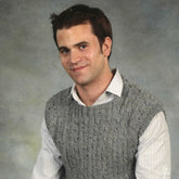 A man with short brown hair and a slight smile is wearing the Men's Cabled Vest by Plymouth Yarn Co, crafted from worsted weight yarn, over a white dress shirt. He is posed in front of a simple, gray background.