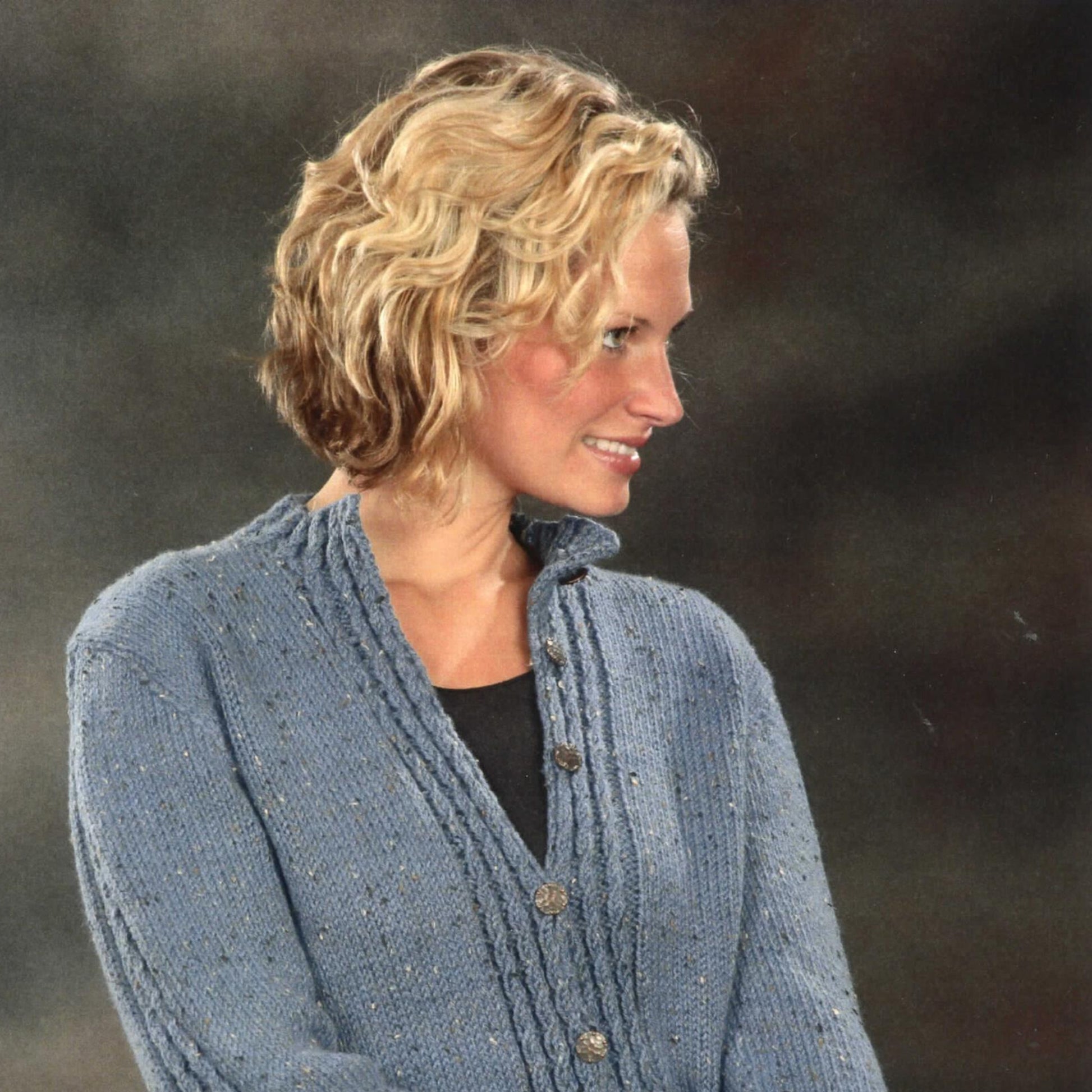 A person with short, wavy blonde hair smiles while looking to their right. They are wearing a blue Plymouth Yarn Co Mock Cable Cardigan knit from soft yarn over a black top. The background is dark and blurred.