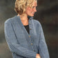 A person with short, wavy blonde hair is wearing a blue Mock Cable Cardigan by Plymouth Yarn Co over a black shirt. The person is facing slightly to the right, smiling, and standing against a greyish, indistinct background.