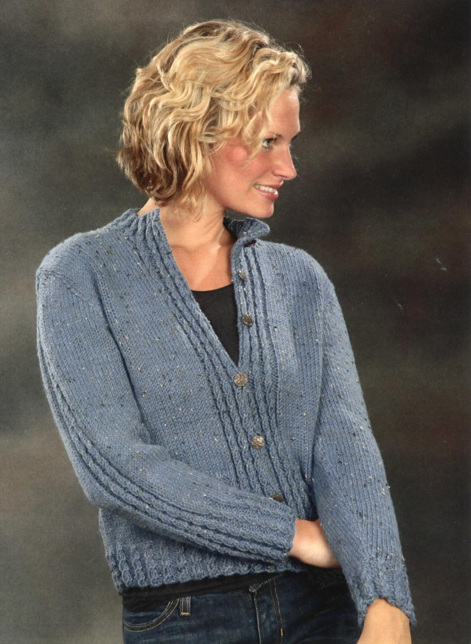 A person with short, wavy blonde hair is wearing a blue Mock Cable Cardigan by Plymouth Yarn Co over a black shirt. The person is facing slightly to the right, smiling, and standing against a greyish, indistinct background.