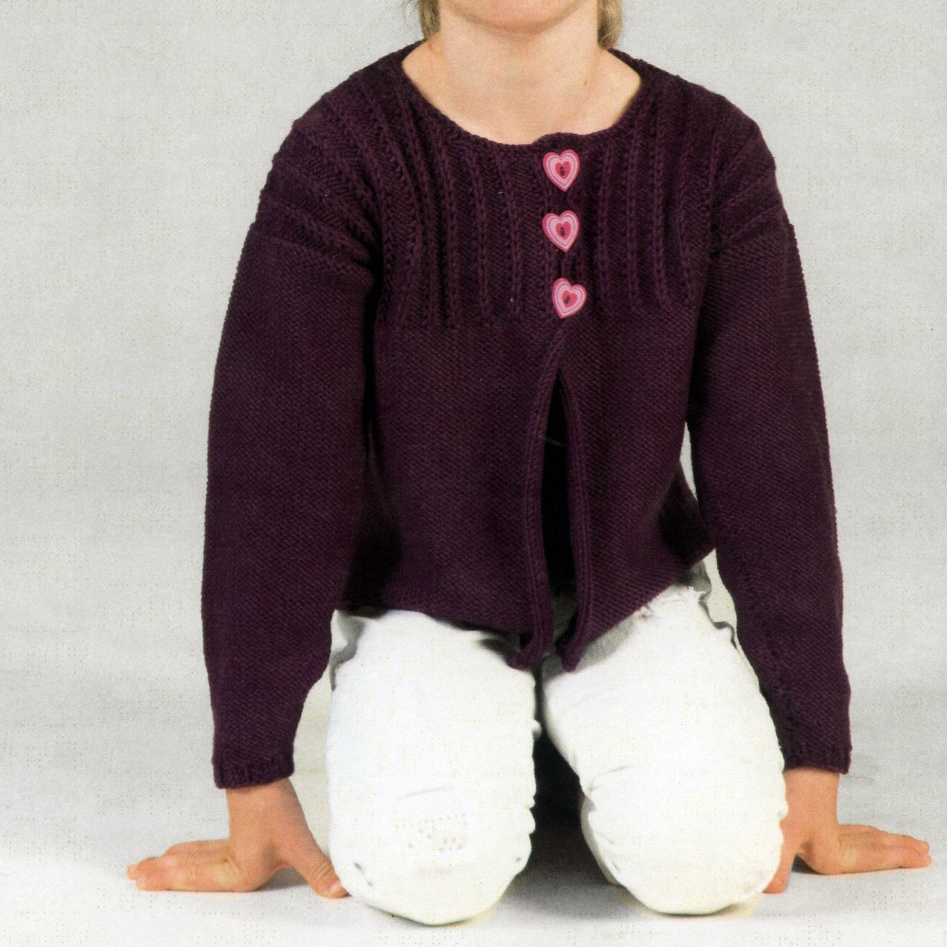 A child is kneeling on the floor, clad in a deep purple Girl's Cardigan from Plymouth Yarn Co made with Worsted Merino Superwash. The cardigan, adorned with heart-shaped pink buttons and featuring a Richelieu Rib pattern on the upper torso and sleeves, is paired with white pants. The child's face remains out of view.