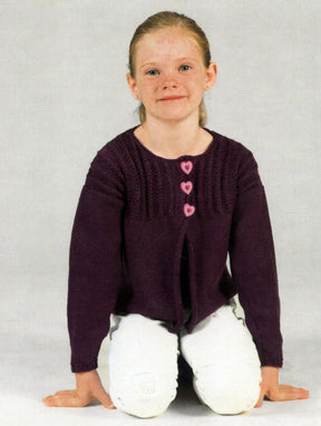 A young girl with light brown hair kneels on the ground, smiling. She is wearing a dark purple girl's cardigan from Plymouth Yarn Co with three pink heart-shaped buttons and white pants. The background is plain light gray.