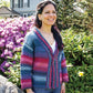 A woman with long dark hair smiles while standing in a garden full of blooming flowers and lush greenery. She is wearing a Patterned Kimono Jacket from Plymouth Yarn Co and jeans. A large, charming house with a gabled roof is visible in the background. The weather appears sunny.