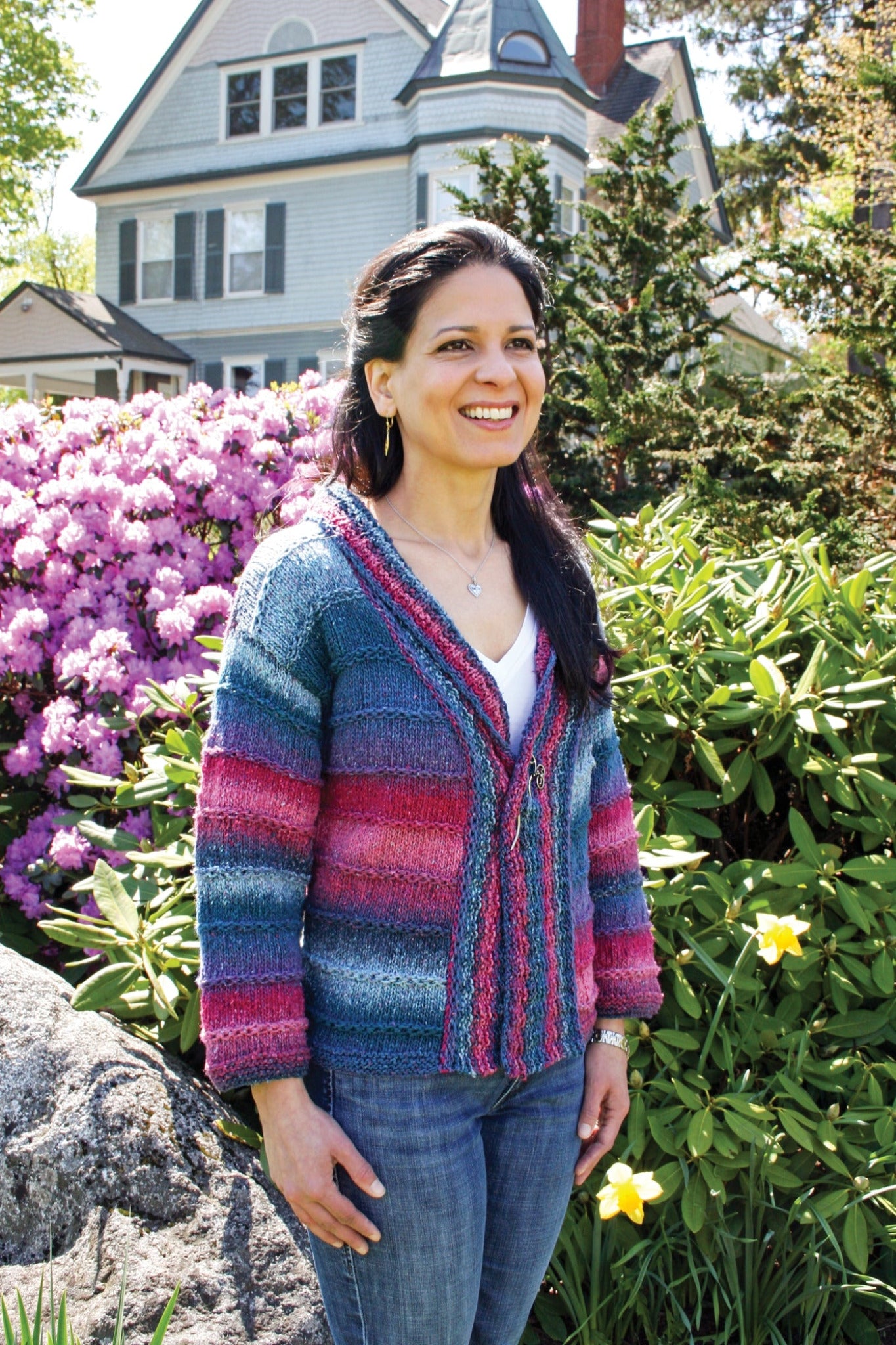 A woman with long dark hair smiles while standing in a garden full of blooming flowers and lush greenery. She is wearing a Patterned Kimono Jacket from Plymouth Yarn Co and jeans. A large, charming house with a gabled roof is visible in the background. The weather appears sunny.