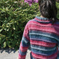 A person with dark hair tied back sits on a concrete surface, facing away from the camera. They wear the Plymouth Yarn Co's Patterned Kimono Jacket, featuring a striped design in shades of pink, blue, and purple that looks like an easy knit. Behind them is a lush green shrub with purple flowers under a clear and sunny sky.