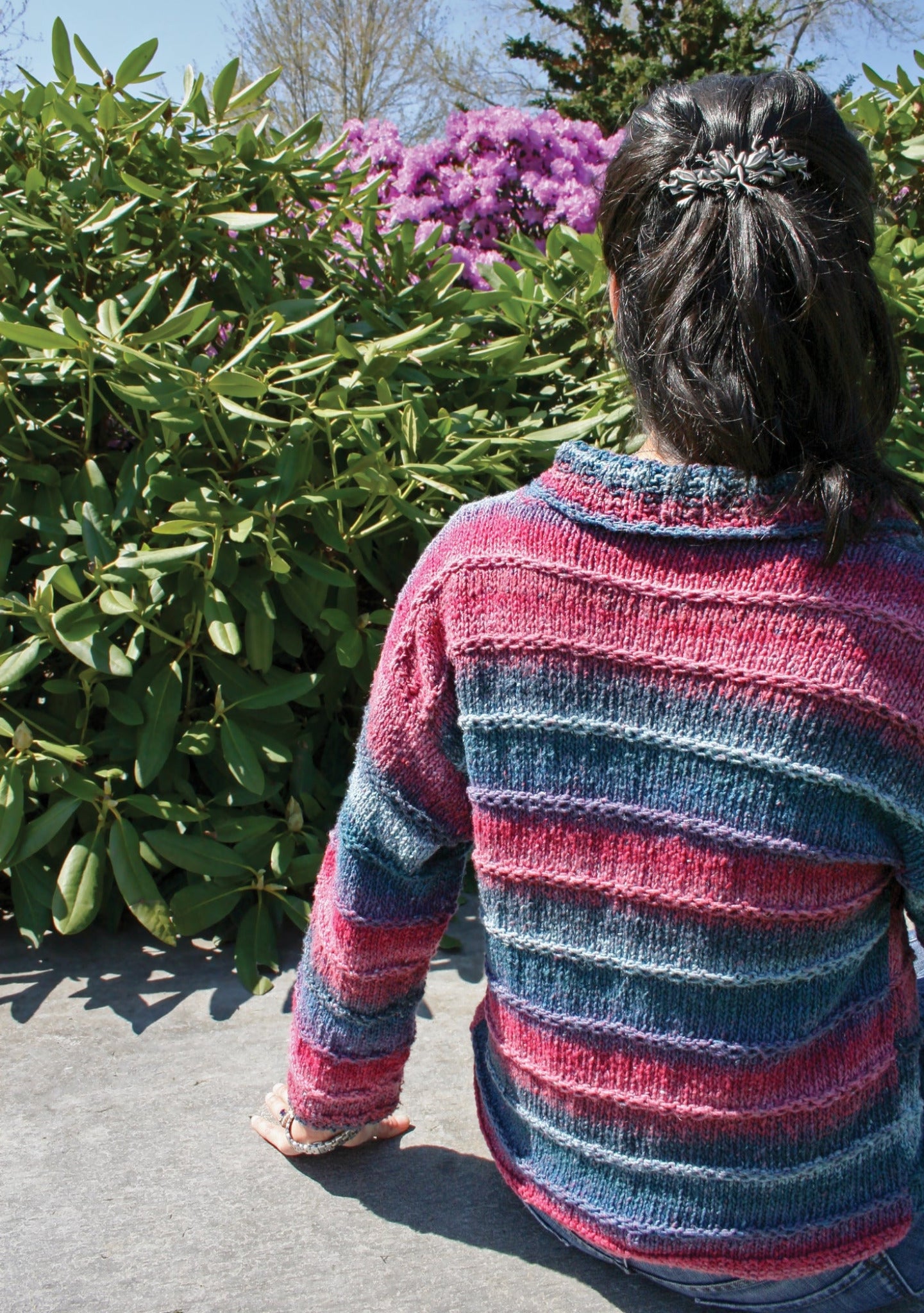 A person with dark hair tied back sits on a concrete surface, facing away from the camera. They wear the Plymouth Yarn Co's Patterned Kimono Jacket, featuring a striped design in shades of pink, blue, and purple that looks like an easy knit. Behind them is a lush green shrub with purple flowers under a clear and sunny sky.