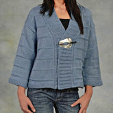 An individual is dressed in a Patterned Kimono Jacket from Plymouth Yarn Co, featuring a light blue hue and textured knit design with wide sleeves. The cardigan is secured at the front with a large, decorative pin. They are also wearing a black top underneath paired with blue jeans, set against a plain gray background.