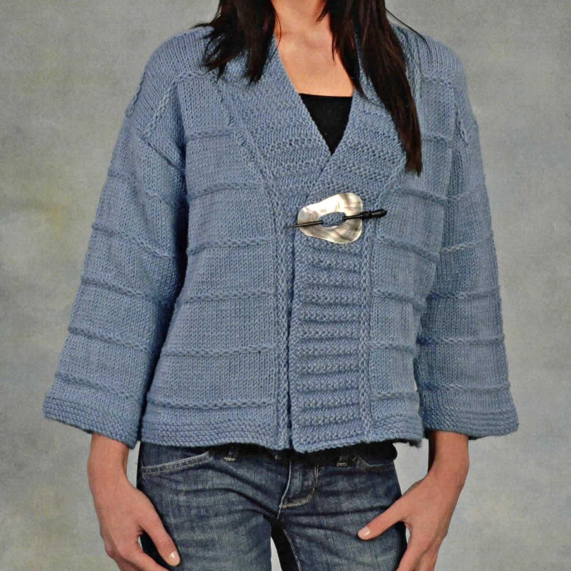 An individual is dressed in a Patterned Kimono Jacket from Plymouth Yarn Co, featuring a light blue hue and textured knit design with wide sleeves. The cardigan is secured at the front with a large, decorative pin. They are also wearing a black top underneath paired with blue jeans, set against a plain gray background.