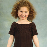 A smiling young girl with curly red hair is wearing a Girl's Smock Dress by Plymouth Yarn Co—an ideal choice for holiday time. She stands against a neutral background with her hands at her sides, exuding the charm associated with this delightful garment.