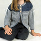 A young girl with long hair wears a "Two Color Child's Jacket" by Plymouth Yarn Co, featuring a blue and white stripe pattern and white buttons. She is sitting cross-legged on the floor, wearing dark jeans. Only her torso and arms are visible in the image.