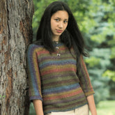 A young woman with long dark hair stands outdoors against a tree trunk, wearing the Kudo Pullover by Plymouth Yarn Co, which features multi-colored stripes and three-quarter length sleeves. The background is filled with greenery, suggesting a park or forest setting.