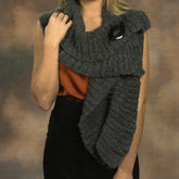 On a cool evening, a person elegantly wraps the Ruffled Wrap Pattern from Plymouth Yarn Co, an asymmetrical gray knitted scarf, over an orange top and black skirt. The scarf features a textured pattern secured with a black pin against the backdrop of a warm, blurred brown gradient.