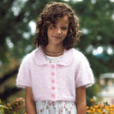 A young girl with curly hair smiles softly while wearing a delicate Angora Girl's Cardigan from Plymouth Yarn Co in light pink, paired with a floral dress. She stands outdoors against a blurred green background, possibly featuring some trees. The knit cardigan is short-sleeved with pink buttons and a Peter Pan collar.