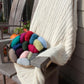 A cozy Plymouth Yarn Co Cabled Afghan drapes over a wooden chair on a patio. A bowl filled with various colorful yarn balls, including red, blue, green, pink, and gray, rests on the chair seat. Sunlight gently illuminates the scene, adding to the warmth and comfort of this cool fall or winter setting.