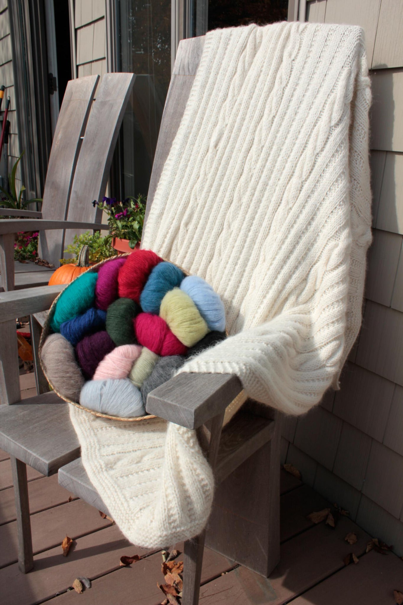 A cozy Plymouth Yarn Co Cabled Afghan drapes over a wooden chair on a patio. A bowl filled with various colorful yarn balls, including red, blue, green, pink, and gray, rests on the chair seat. Sunlight gently illuminates the scene, adding to the warmth and comfort of this cool fall or winter setting.